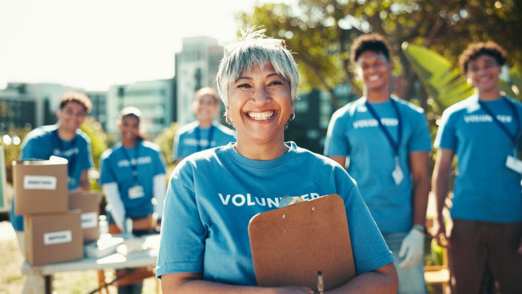 Volunteer smiling woman with clipboard at outdoor community event, diverse team in background.