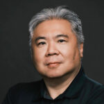 Senior Asian man with gray hair in professional portrait, dark background, focused expression.