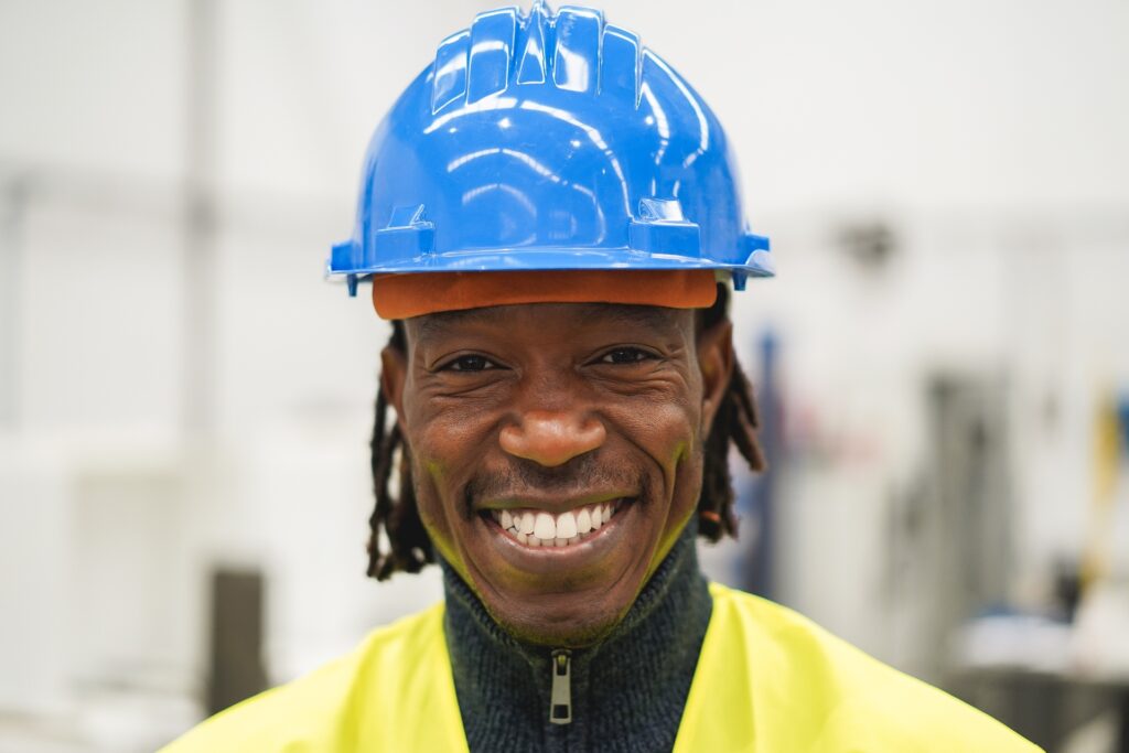 Smiling male industrial worker wearing a blue safety helmet and yellow high-visibility vest.
