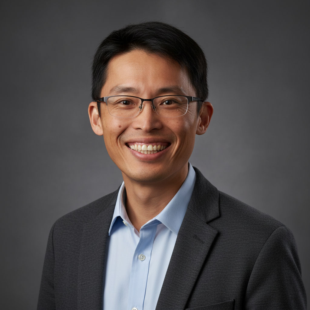 Asian professional man in glasses smiling, wearing a suit against gray background.