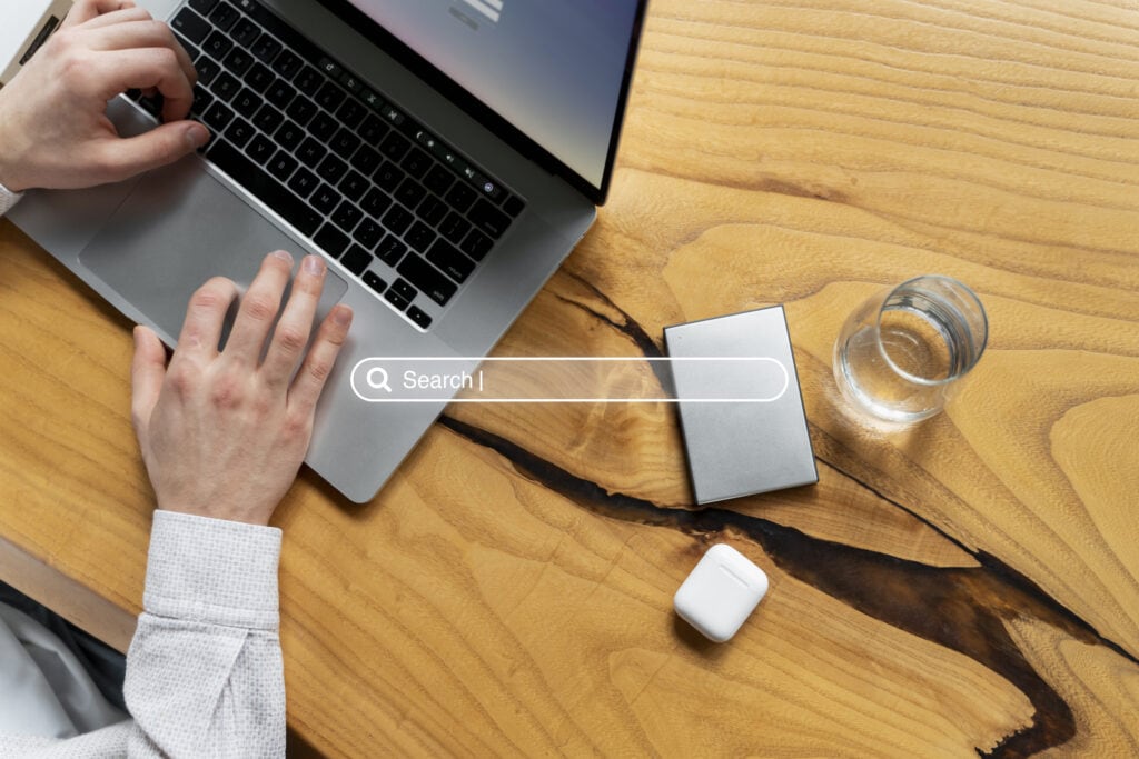 Search laptop on wooden desk with wireless earbuds and glass of water, modern workspace, tech accessories, Concentrus branding.