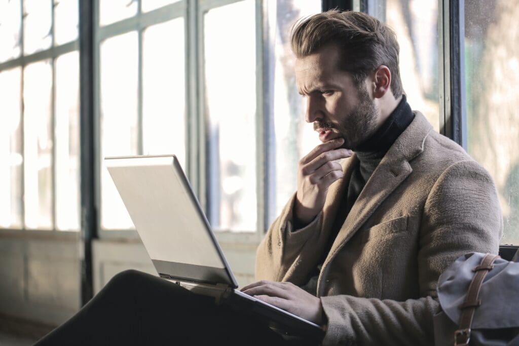 Confused man working on a laptop, focusing intensely, in a modern bright office setting.