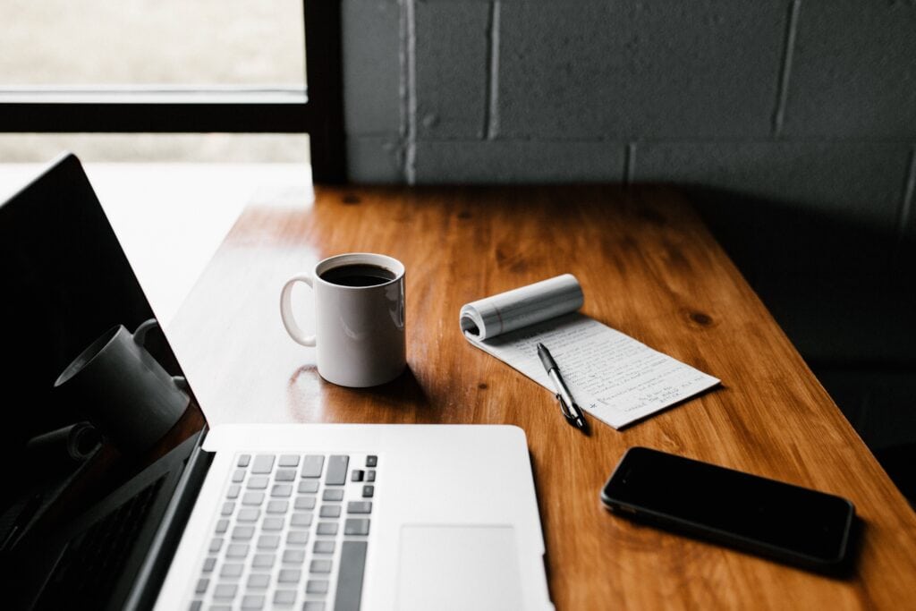 Coffee mug on wooden desk with laptop, notepad, pen, and smartphone in a modern office setting.