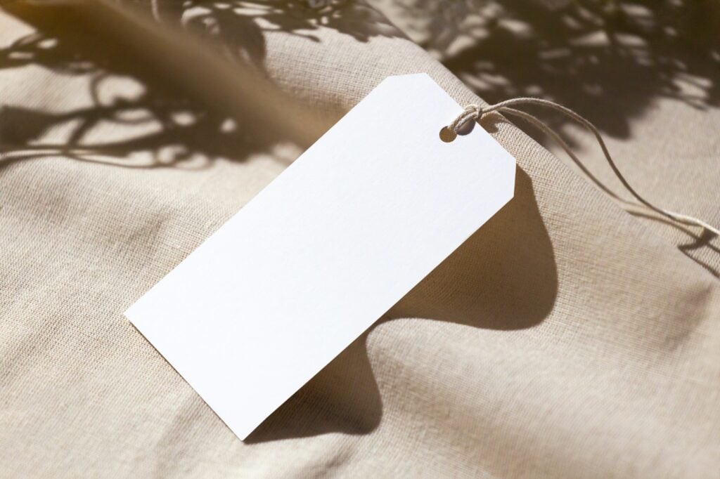 Blank white tag on beige fabric with shadow and string, modern minimalistic design.