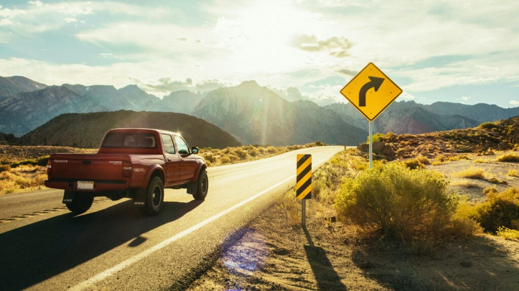 Red pickup truck driving on desert highway with winding road sign in scenic mountain landscape.