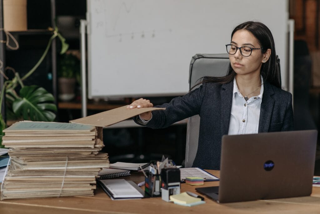 Overwhelmed woman managing paperwork in an office workspace with files and a laptop.