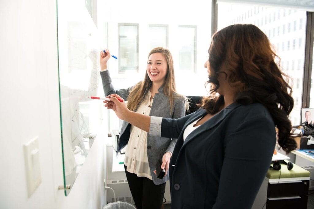 Collaborative women discussing data analysis at office whiteboard, teamwork, business strategy.