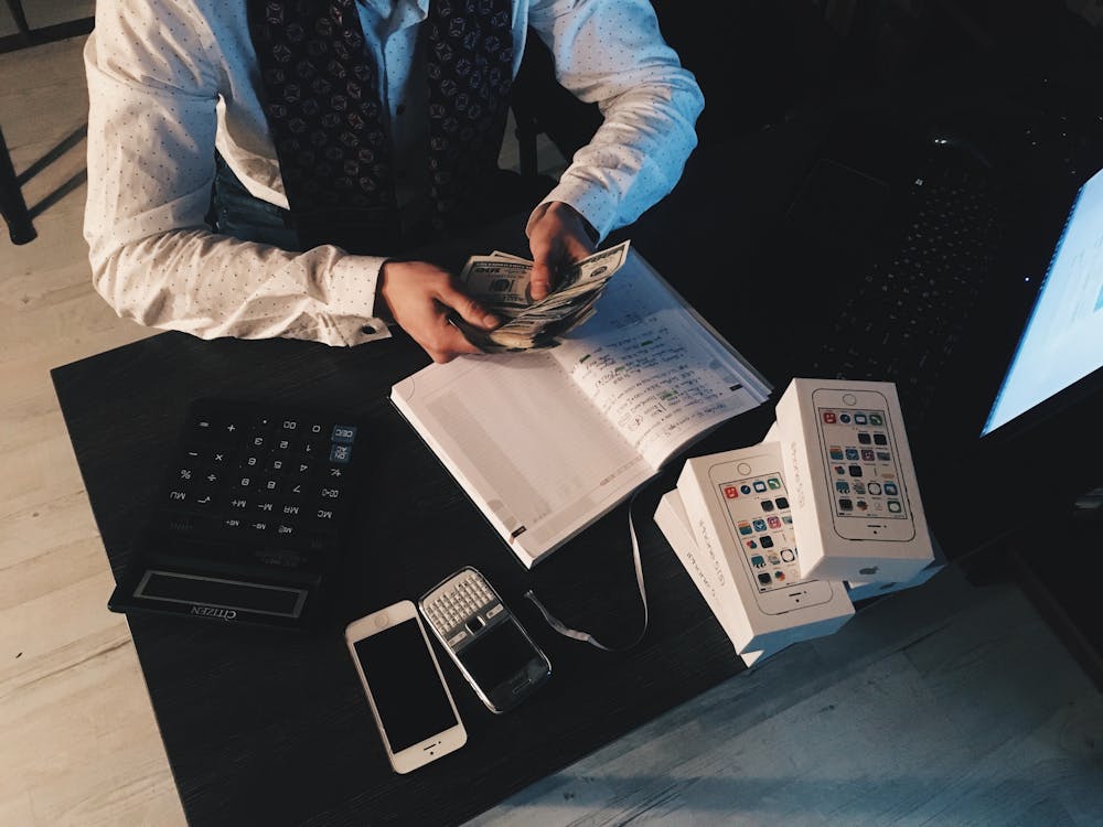 Stressed businessman counting cash with calculator, phone, and unopened smartphone boxes on desk.
