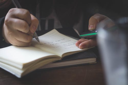 Hand writing in notebook with pen and phone on table, focused and organized workspace.