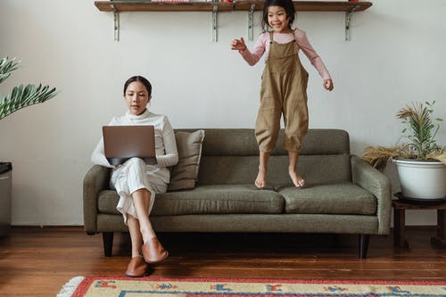 Young girl jumping on sofa with woman working on laptop at home, family, comfort, relaxation, modern living.