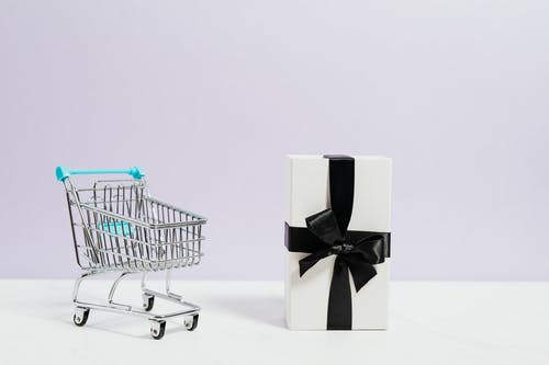 Miniature shopping cart with a gift box wrapped in black ribbon on a white background.