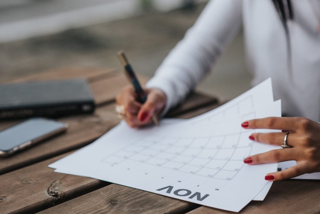 Close-up of woman writing on November calendar with pen at wooden desk with notebook and phone in background.