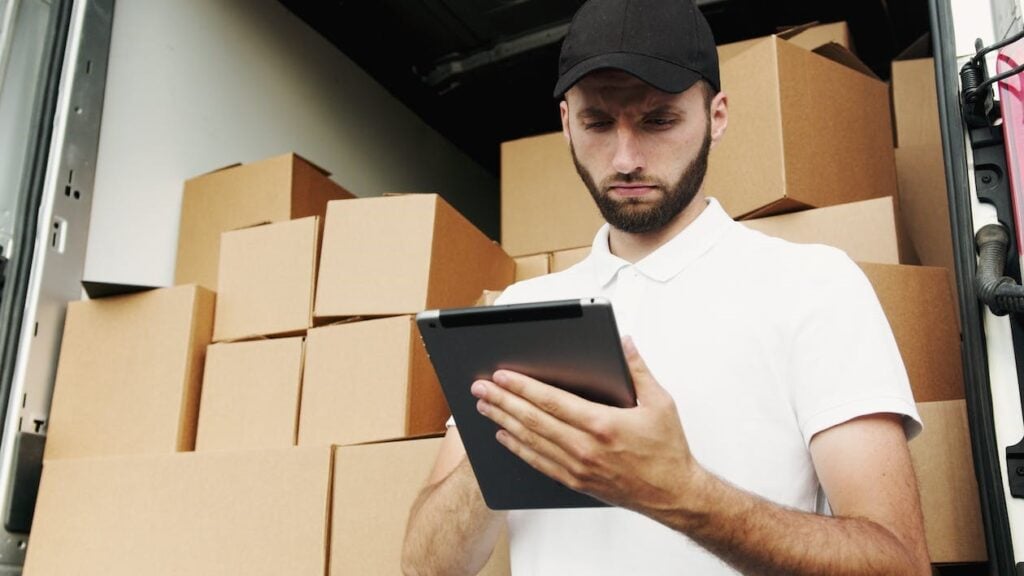 1. Delivery driver checking tablet among packed boxes at warehouse.